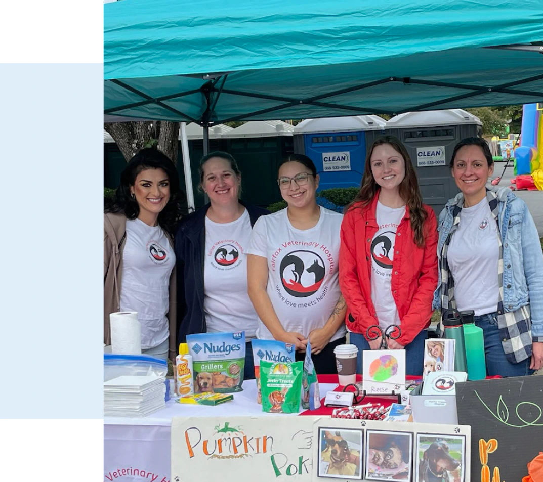 Contact_Us_Join_Our_Team_Rectangle Five smiling women stand behind a booth under a teal canopy. They wear matching shirts with a veterinary logo. The table displays pet treats, flyers, and a sign for “Pumpkin Poke.” Portable toilets are visible in the background.
