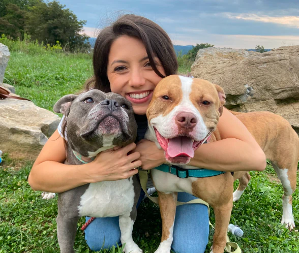 Mobile_MTD_Gabriella A smiling woman kneels outdoors, hugging two happy dogs—one gray and white, the other brown and white—against a backdrop of grass, rocks, and trees under a cloudy sky.