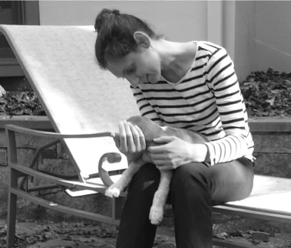 Mobile_MTD_Lauren A woman in a striped shirt sits on an outdoor lounge chair, smiling and gently holding a small dog on her lap. The setting appears to be a patio with leaves scattered on the ground.