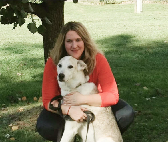 Mobile_MTD_Melissa A woman with long blonde hair in an orange top is kneeling on grass, hugging a large light-colored dog while smiling at the camera. There is a tree and green lawn in the background.