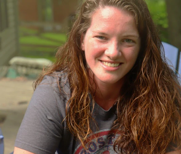 A woman with long, wavy brown hair and a gray T-shirt smiles while sitting outdoors on a patio with greenery and outdoor furniture in the background.