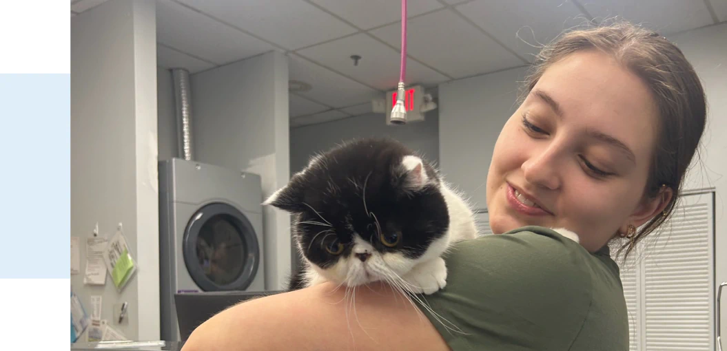 Services_Acupuncture&_Rectangle A young woman with a slight smile holds a black and white Persian cat over her shoulder in a brightly lit room with a laundry machine and cabinets in the background.